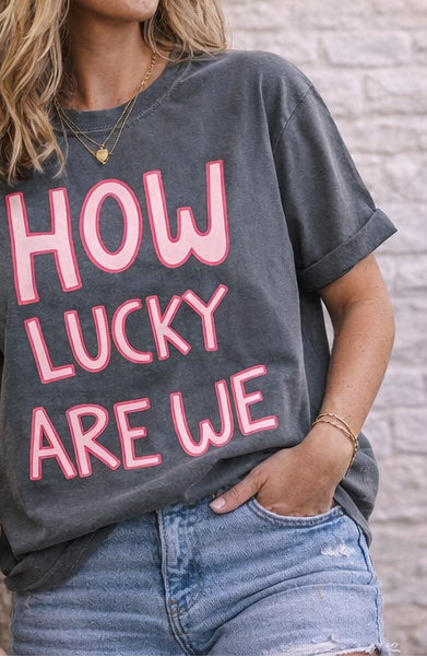 Gray t-shirt with pink text 'HOW LUCKY ARE WE' on a textured surface.