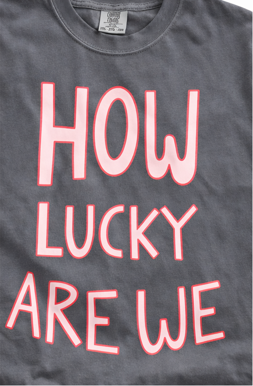 Gray t-shirt with 'HOW LUCKY ARE WE' text in pink, displayed on a hanger and worn by a person.