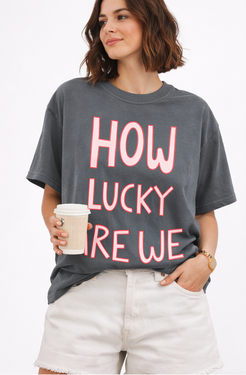 Gray t-shirt with 'HOW LUCKY ARE WE' text in pink, displayed on a hanger and worn by a person.